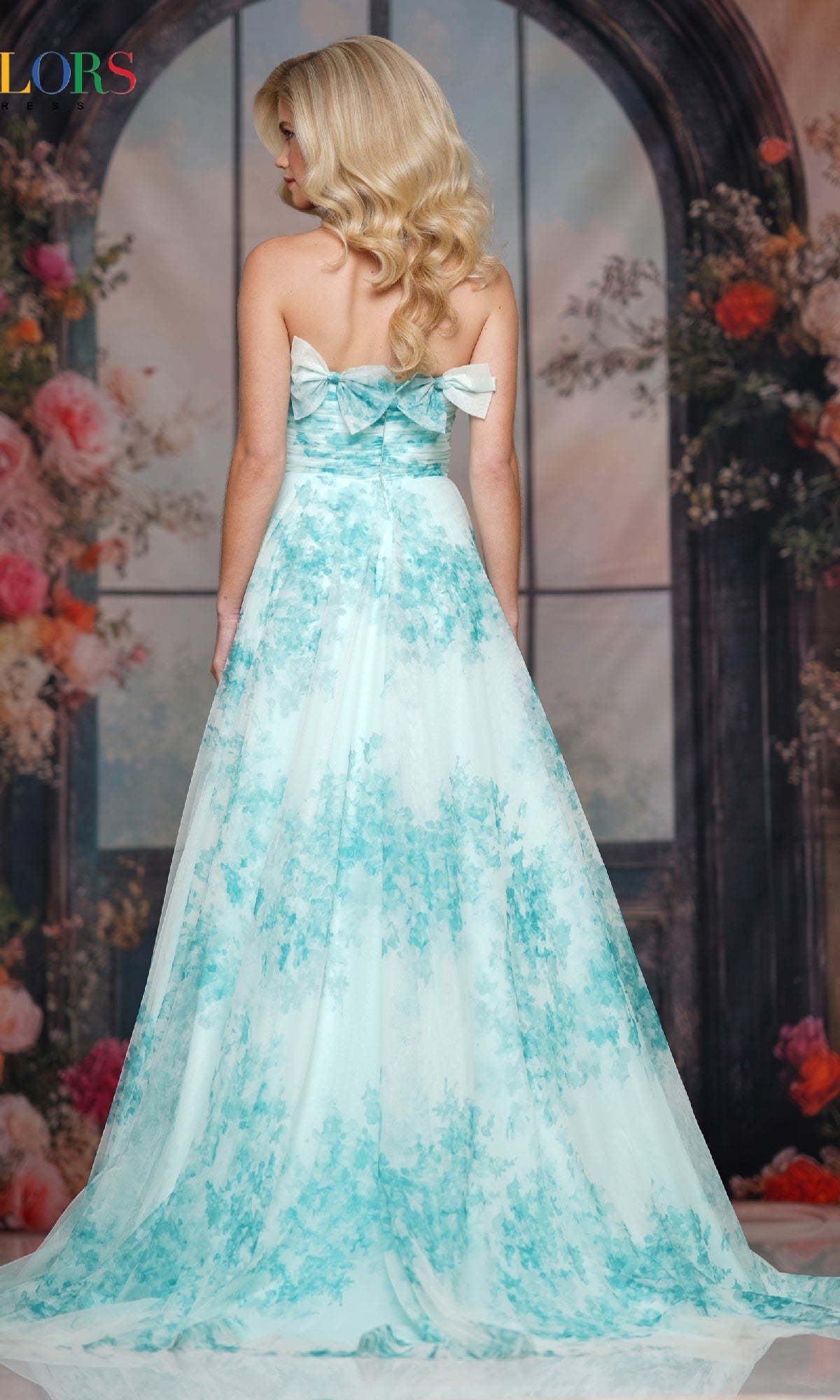 A woman with blonde hair faces away in front of a floral and arch window backdrop, wearing Colors Dress 3811—a strapless long prom dress with an a-line skirt.